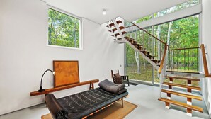 A modern interior with a sofa, a wooden bench, a staircase, and large windows offering a forest view. - Modern home sited on a lush wood acre, stunning pool, covered deck, outdoor... (East Hampton)