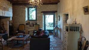 A rustic room with a stone fireplace, wooden furniture, a chandelier, and a view through a window. - Beautiful Lotoise house, located in a typical village. Large garden and swimming pool. (Le Vignon-en-Quercy)