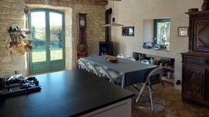 A rustic kitchen with a stone wall, wooden beams, a large window, a dining table, chairs, a stove, and various kitchen utensils. - Beautiful Lotoise house, located in a typical village. Large garden and swimming pool. (Le Vignon-en-Quercy)