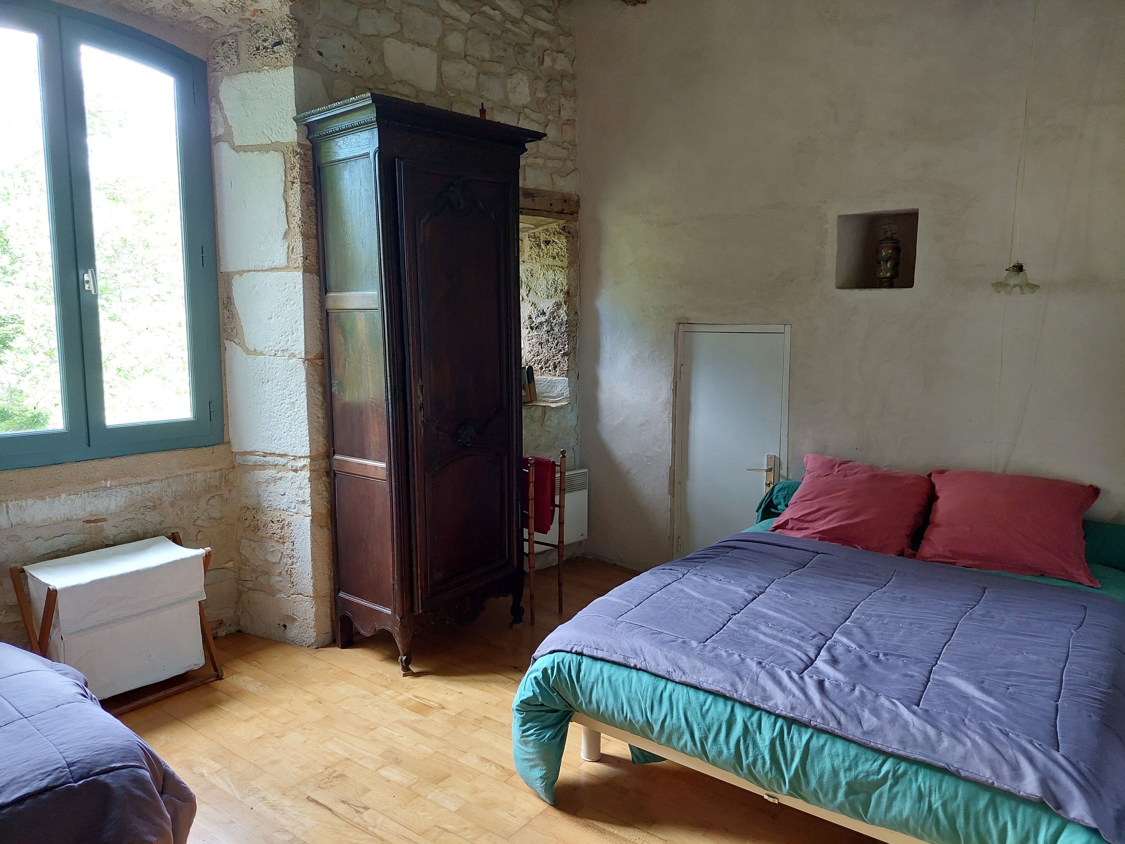 Un dormitorio con un armario de madera, una cama con colcha azul y verde, y una ventana con vista a follaje.
