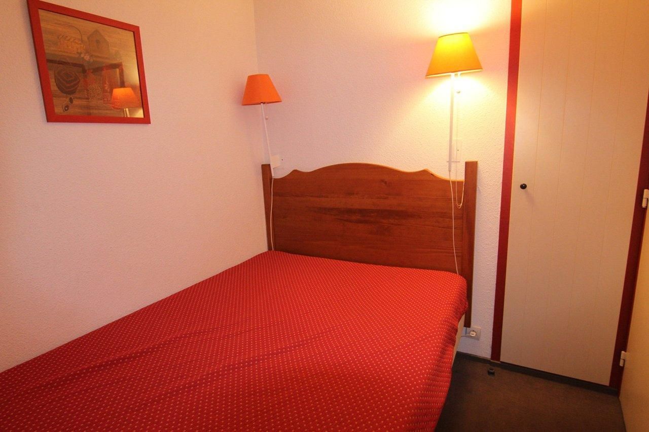 Un lit simple avec une tête de lit en bois, une couvre-lit rouge, deux lampes fixées au mur et un tableau encadré accroché au mur.