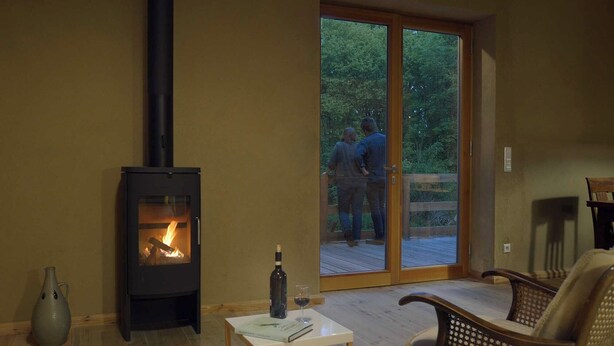 A cozy room with a wood-burning stove, a glass door leading to an outdoor deck, and a small table with a bottle and glass. - Pure tranquillity: vacation apartment close to nature in Unkengrund (Nordwestuckermark)