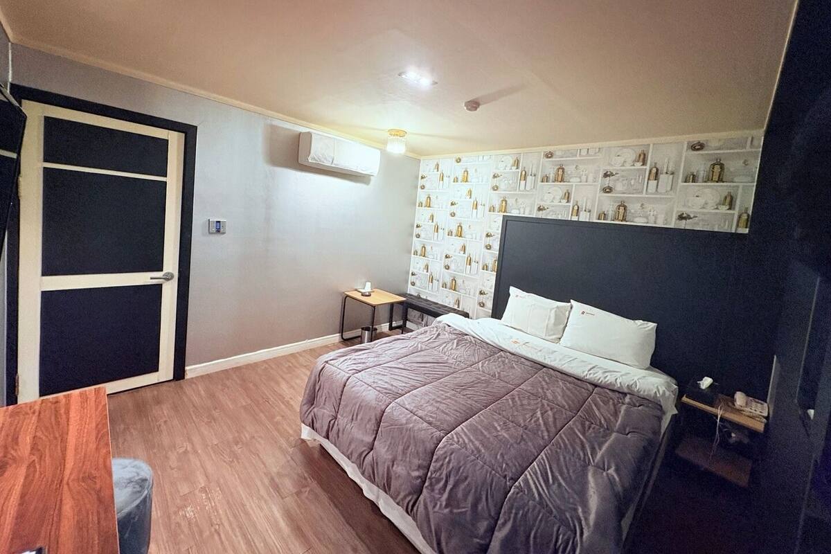 A bedroom with a bed, a small bedside table, a wall-mounted shelf with decorative items, and a door.