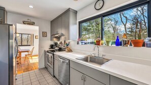 A modern kitchen with stainless steel appliances, a large window with a view of trees, and a dining area with wooden chairs.