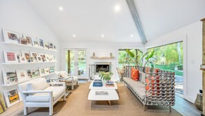 A spacious living room with a fireplace, a bookshelf filled with framed photos, a white sofa, and a coffee table with books.
