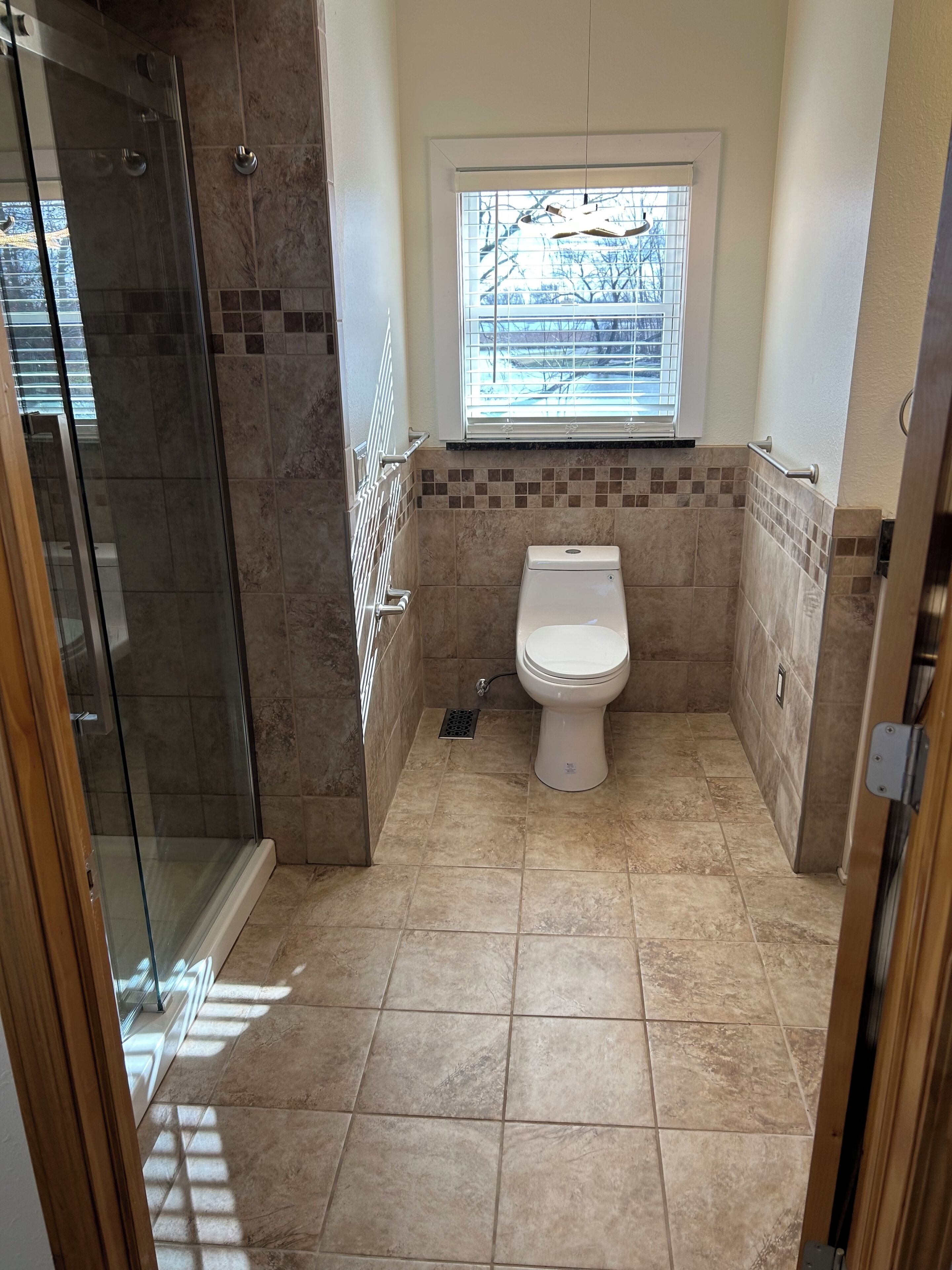 A bathroom with a glass shower enclosure, a toilet, and a window with blinds.