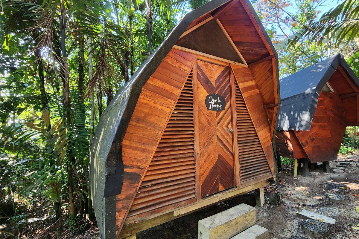 Two wooden cabins with a chevron design, labeled "Cajuna Amigos," set in a forested area with a dirt ground.