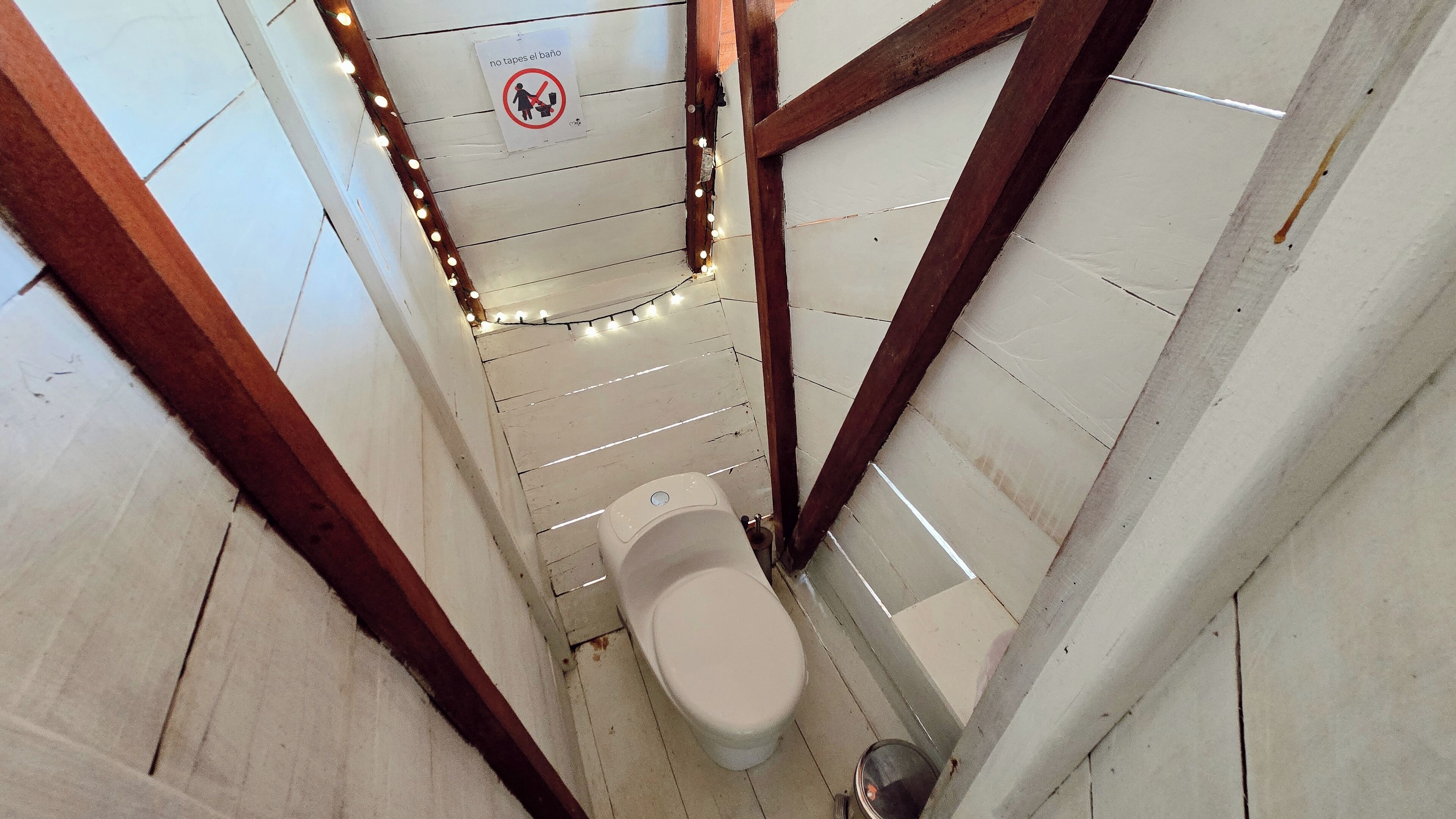 Une petite salle de bain carrelée, avec des toilettes, un panneau indiquant qu’il est interdit de se baigner, et des guirlandes lumineuses accrochées au plafond.