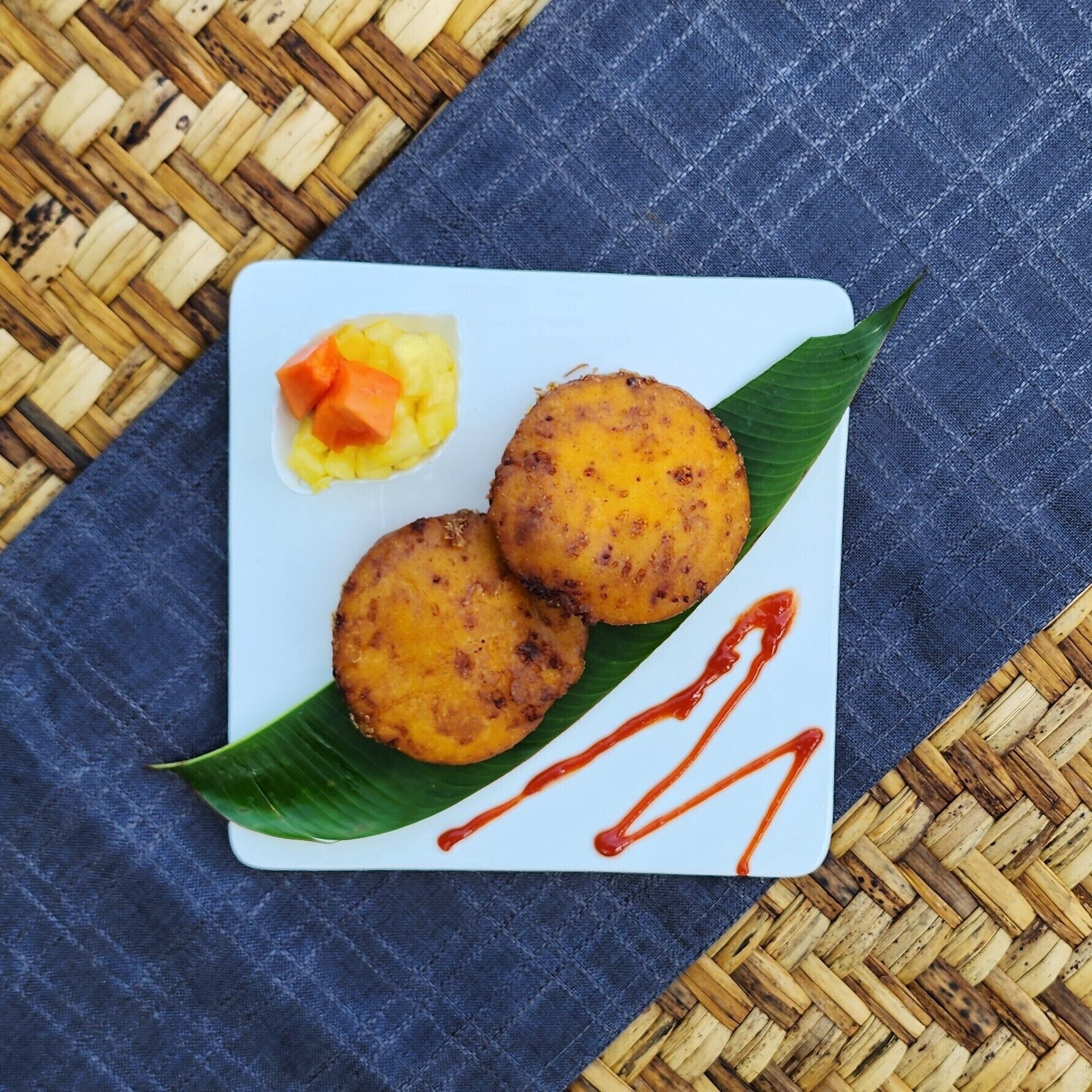 Dos porciones redondas y doradas, fritas, sobre un plato blanco con una hoja verde, acompañadas de una pequeña porción de verduras amarillas y anaranjadas en cubos y salsa roja.