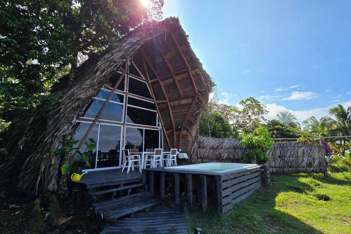 A thatched-roof structure with a large glass window and a wooden deck with chairs.