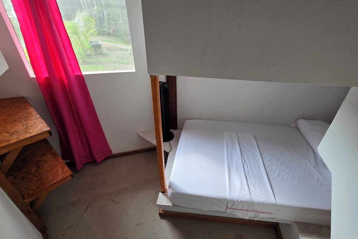 A small, simple room with a bed, a wooden table, and a window with a curtain.