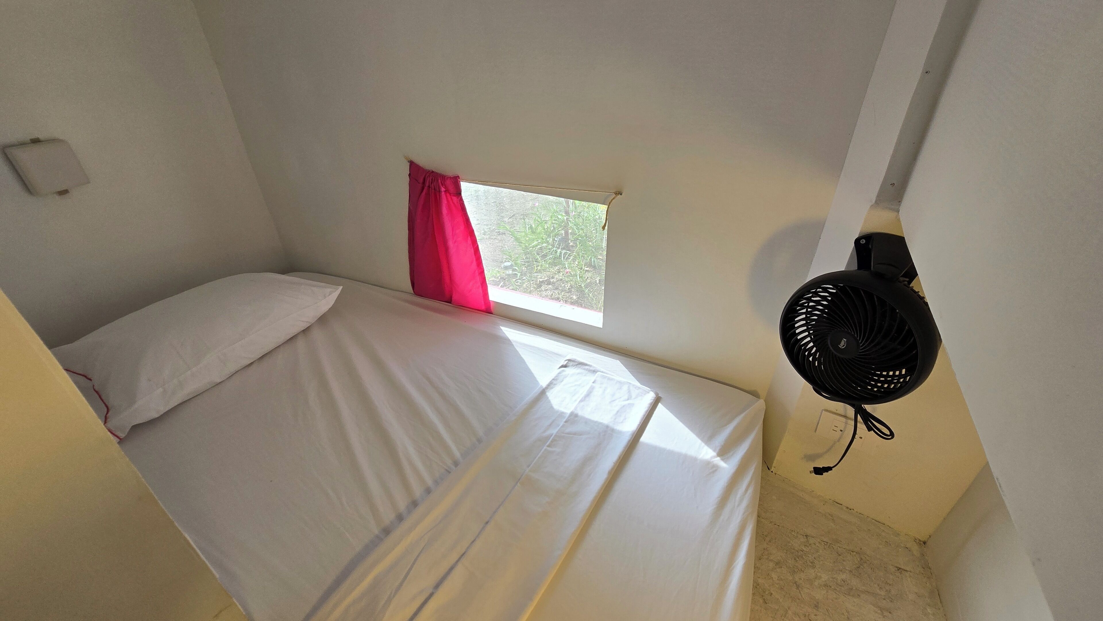 A small, single-bed room with a window, a fan, and a power outlet on the wall.