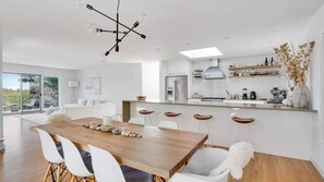 Una cocina moderna con una isla central, armarios blancos y una zona de comedor con una mesa de madera y sillas blancas.