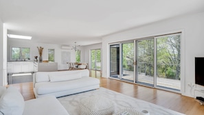 Una sala moderna con un sofá, un comedor con mesa y sillas, una cocina con armarios blancos, y amplios puertas corredizas de vidrio a una terraza exterior.