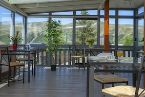 A sunlit outdoor dining area with wooden tables and chairs, a potted plant, and a view of the countryside.