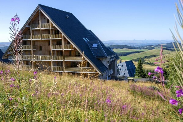 A wooden chalet with a sloped roof, multiple balconies, and a scenic view of rolling hills.