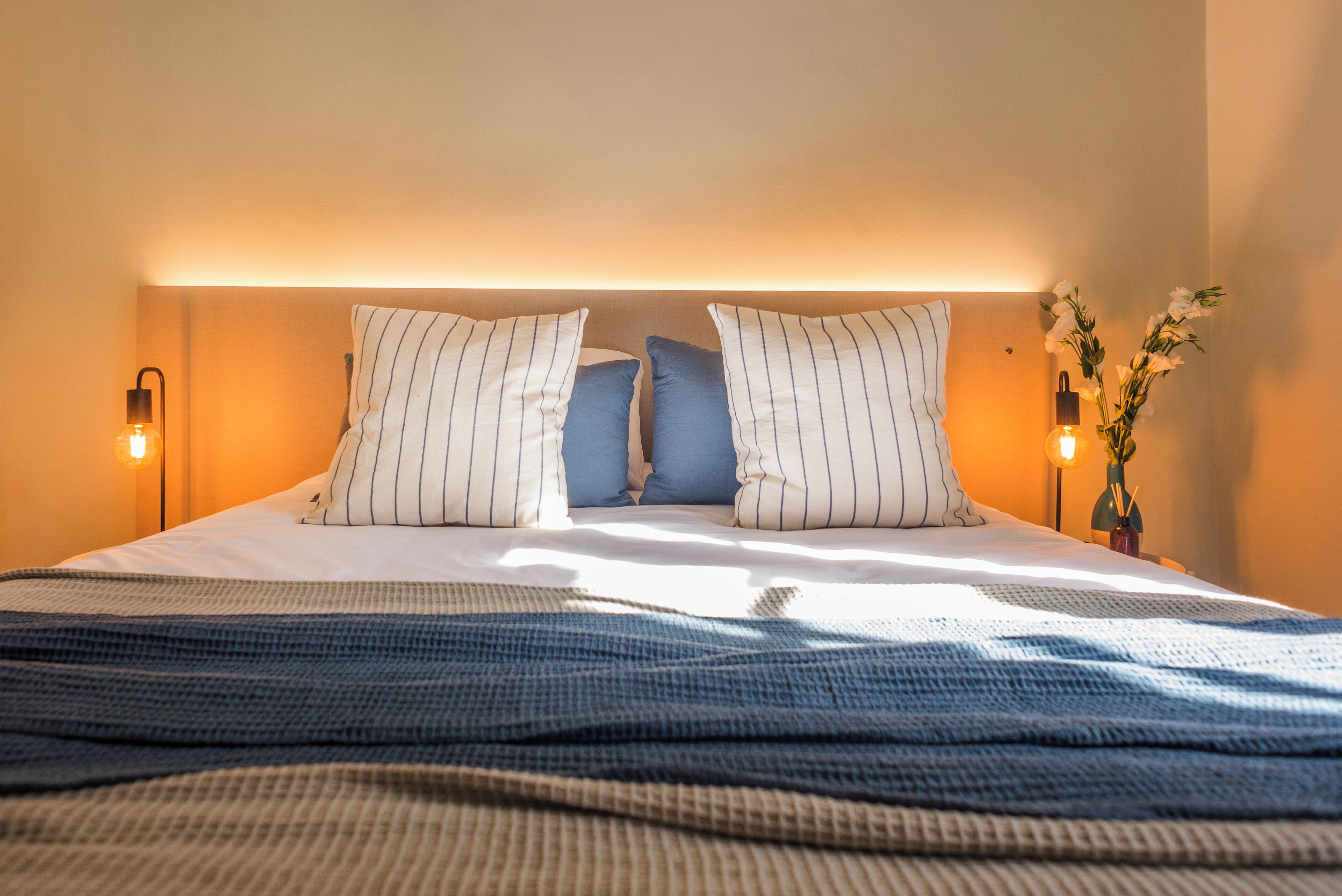 Una cama bien tendida con almohadillas a rayas, una colcha azul y dos lámparas fijadas a la pared.