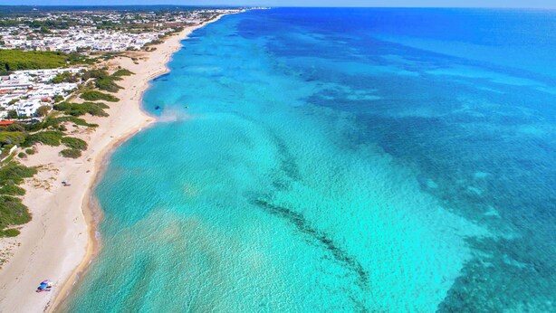 Luftaufnahme einer Küstenlandschaft mit Strand, klarem türkisblauem Wasser und einer Wohngegend mit Häusern und Grünflächen.