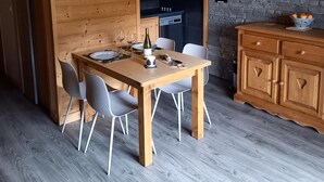 Uma mesa de jantar de madeira com cadeiras, um relógio na parede e um muro de pedra ao fundo.