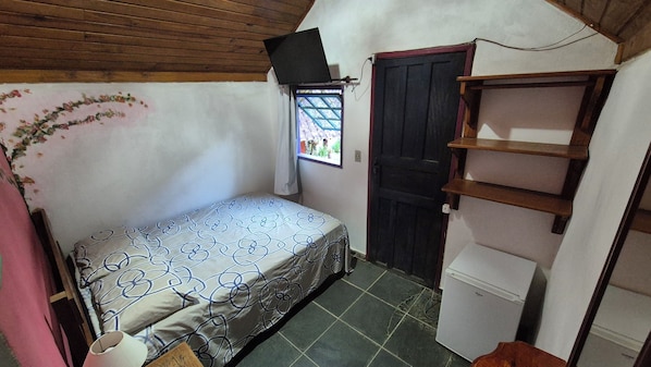 A small, compact room with a bed, a door, a window, and a shelf unit. - Brilho do Sol (Nova Friburgo)