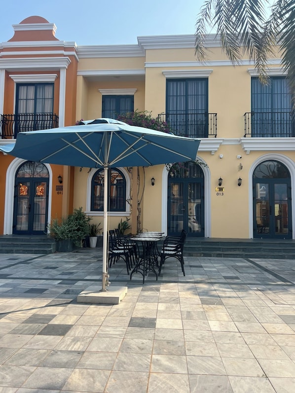 A patio area with a table and chairs under a blue umbrella, surrounded by a tiled floor and potted plants.