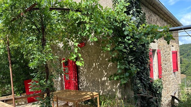 A rustic stone building with a wooden table and red shutters, surrounded by greenery and vines. - ARDECHE COUNTRY GITE OPEN ALL YEAR ROUND (Silhac)
