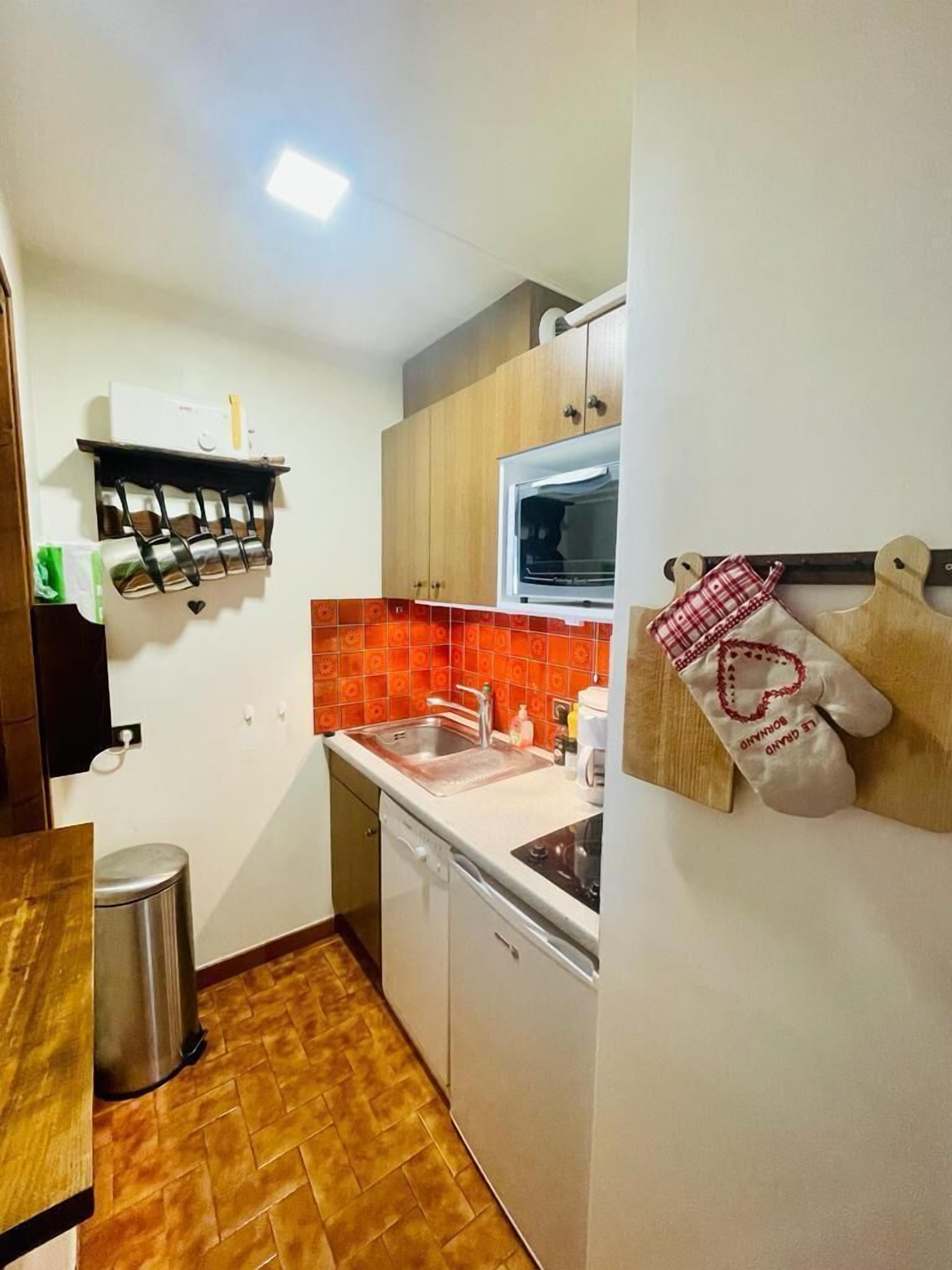 Une cuisine compacte équipée d’un évier, d’une cuisinière et d’un four. Un égouttoir à vaisselle est placé au-dessus de l’évier et une poubelle se trouve dans un coin.