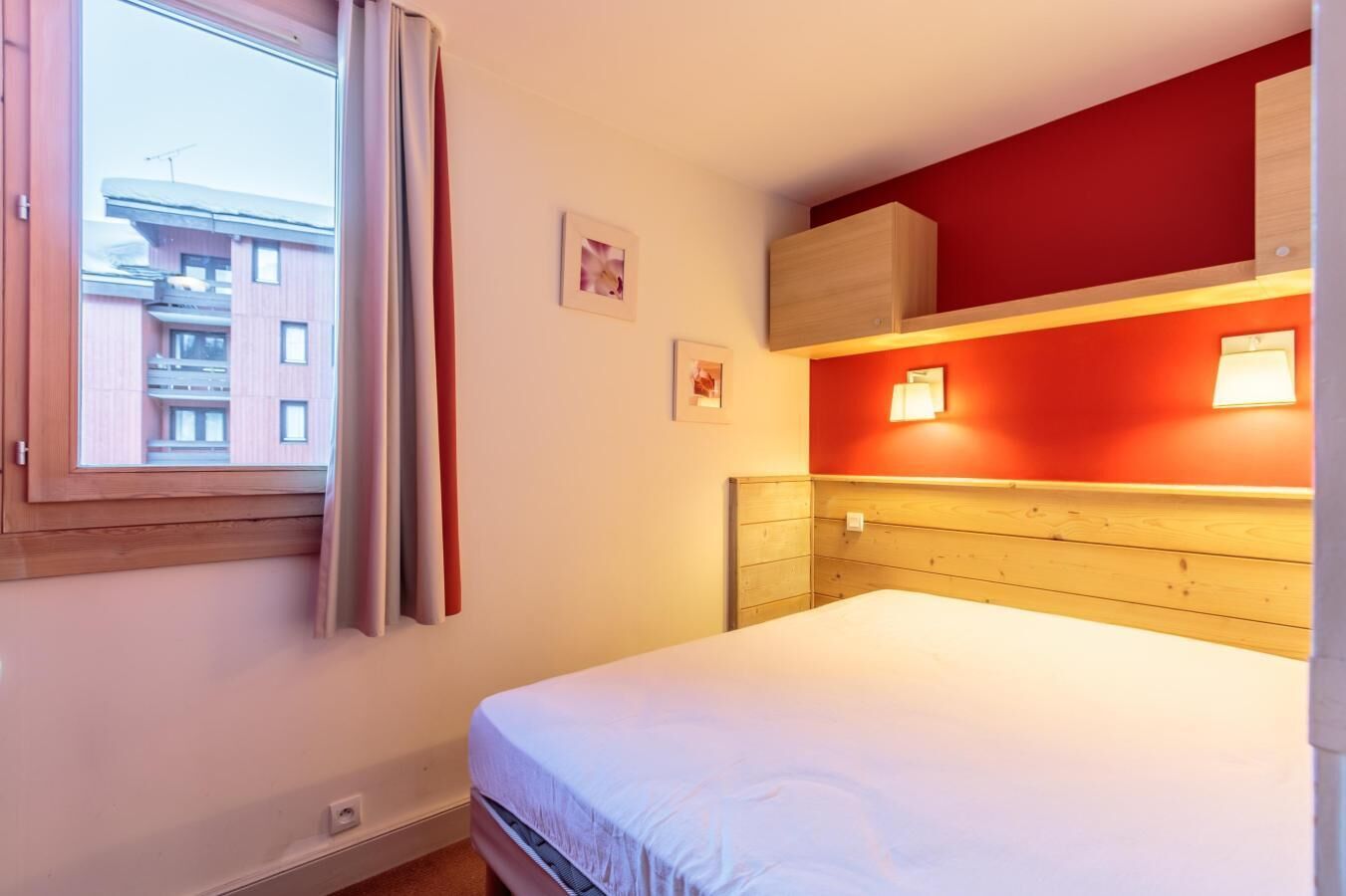 Een hotelkamer met een bed, een raam met gordijnen, een rode muur en een houten hoofdbord.