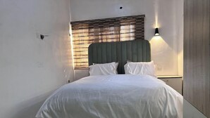Una cama bien hecha con cabecera, una lámpara de noche y una ventana con persianas.