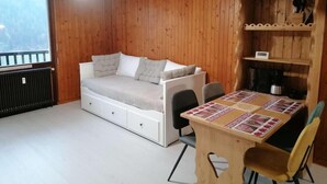 Een gezellige kamer met een houten wandpaneel, een bed met opbergladen, een eettafel met een tafelkleed met patroon, en een inbouwkastje met een waterkoker.