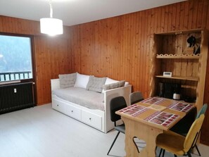 Ein gemütliches Zimmer mit einer holzverkleideten Wand, einem Bett mit Stauraum, einem Esstisch mit gemustertem Tischlauf und einer integrierten Ablage mit Wasserkocher.