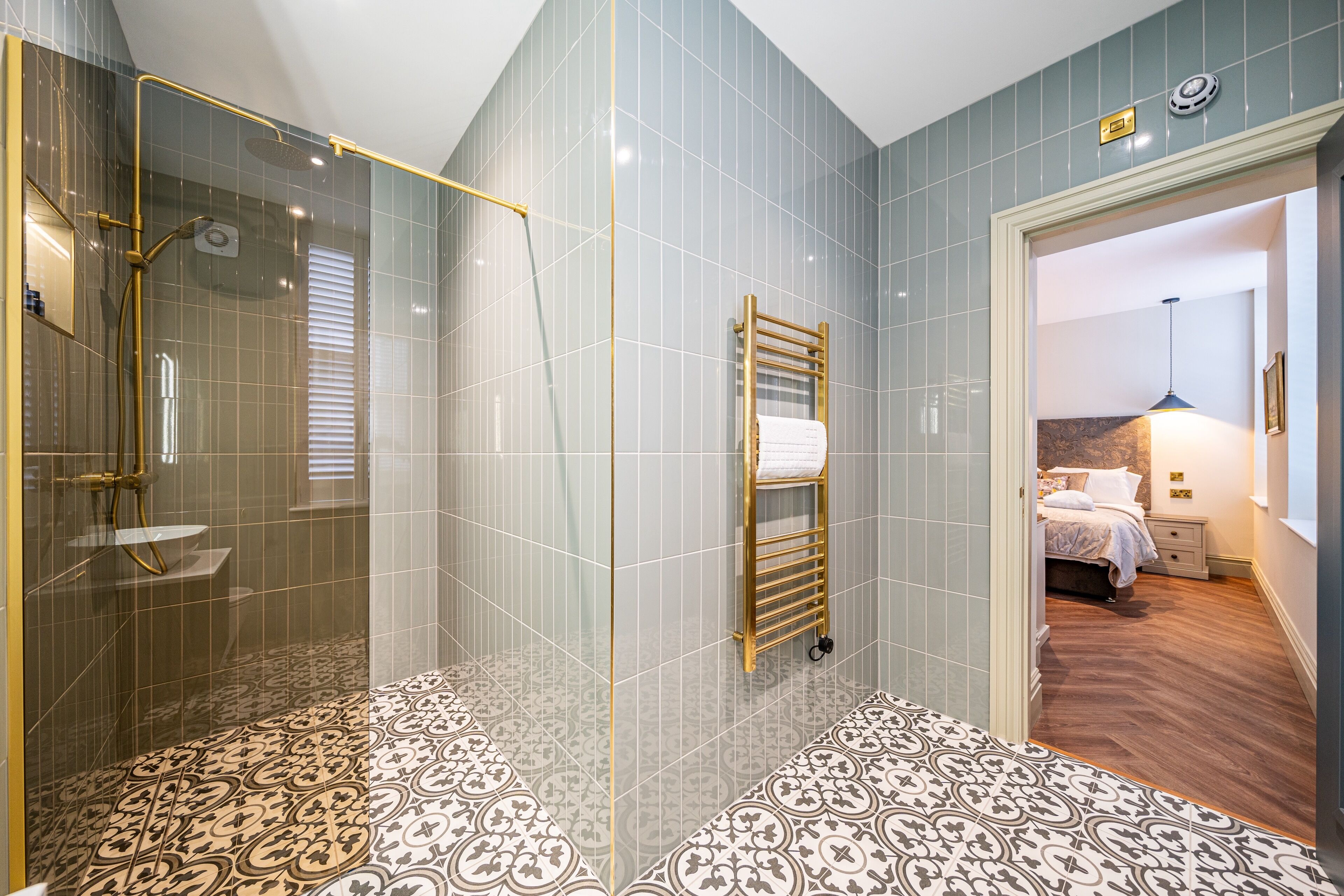 A modern bathroom with a glass-enclosed shower, patterned floor tiles, and a visible bedroom with a bed and nightstand.