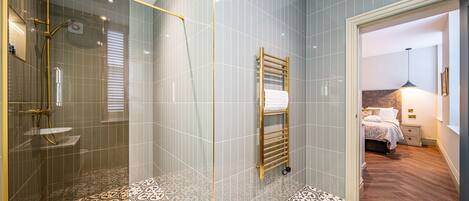 A modern bathroom with a glass-enclosed shower, patterned floor tiles, and a visible bedroom with a bed and nightstand.
