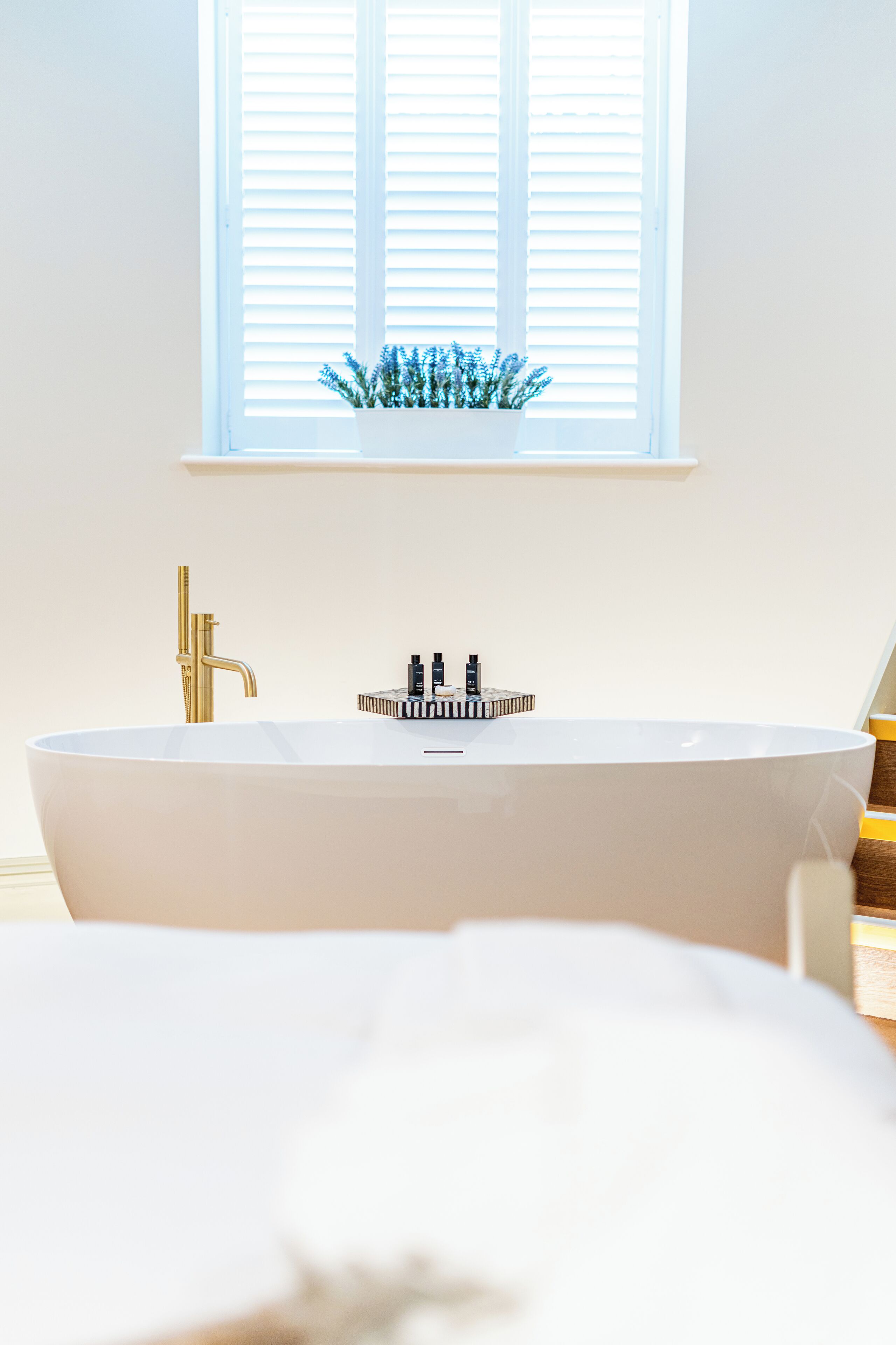 A modern bathroom with a freestanding bathtub, a gold faucet, and a window with blinds.