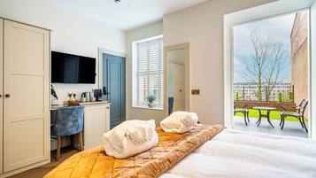 A hotel room with a bed, a desk, a TV, a window with blinds, and a balcony with chairs.