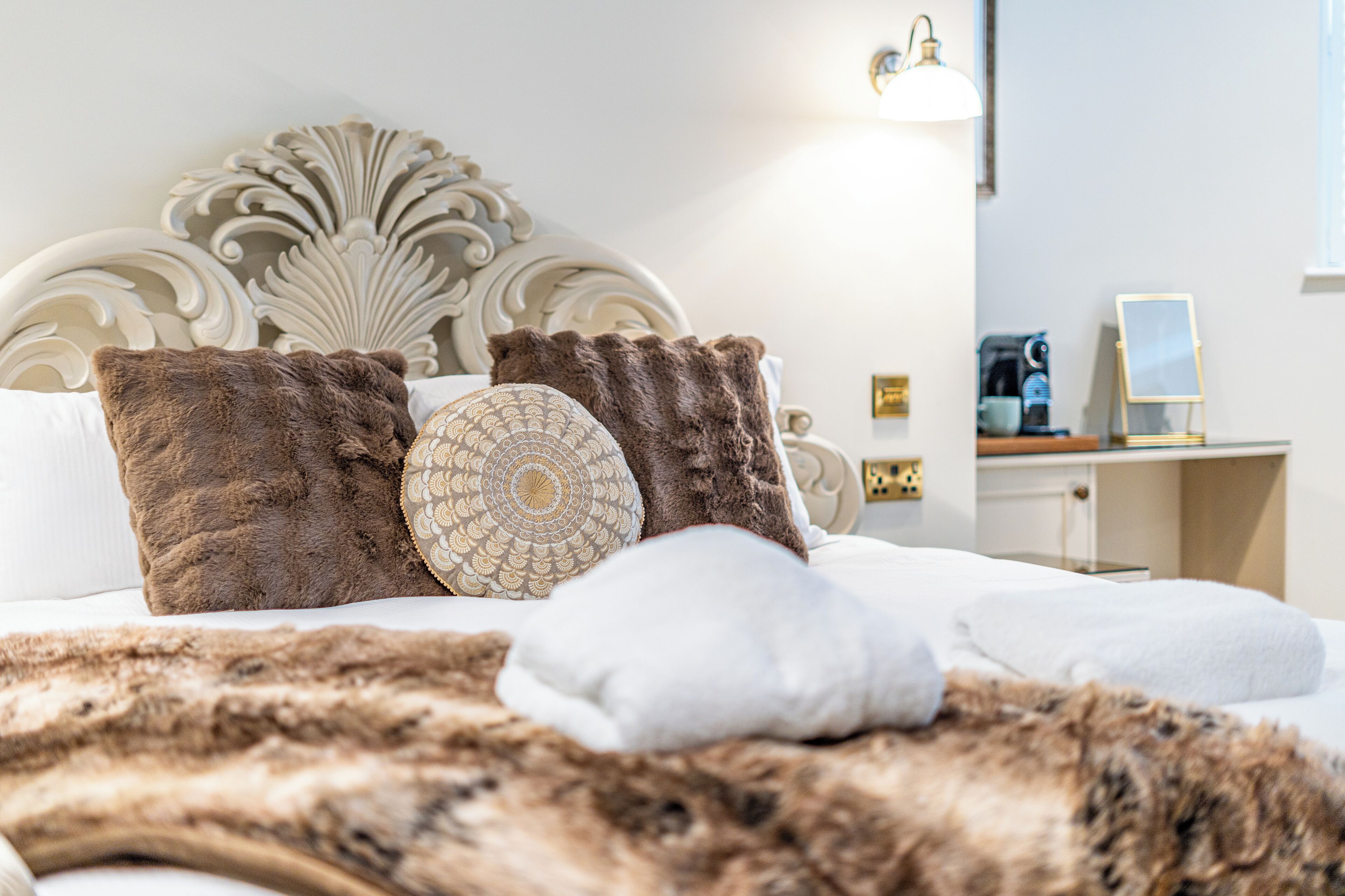 A neatly made bed with a fluffy brown blanket, a decorative headboard, and a bedside table with a lamp and a coffee maker.