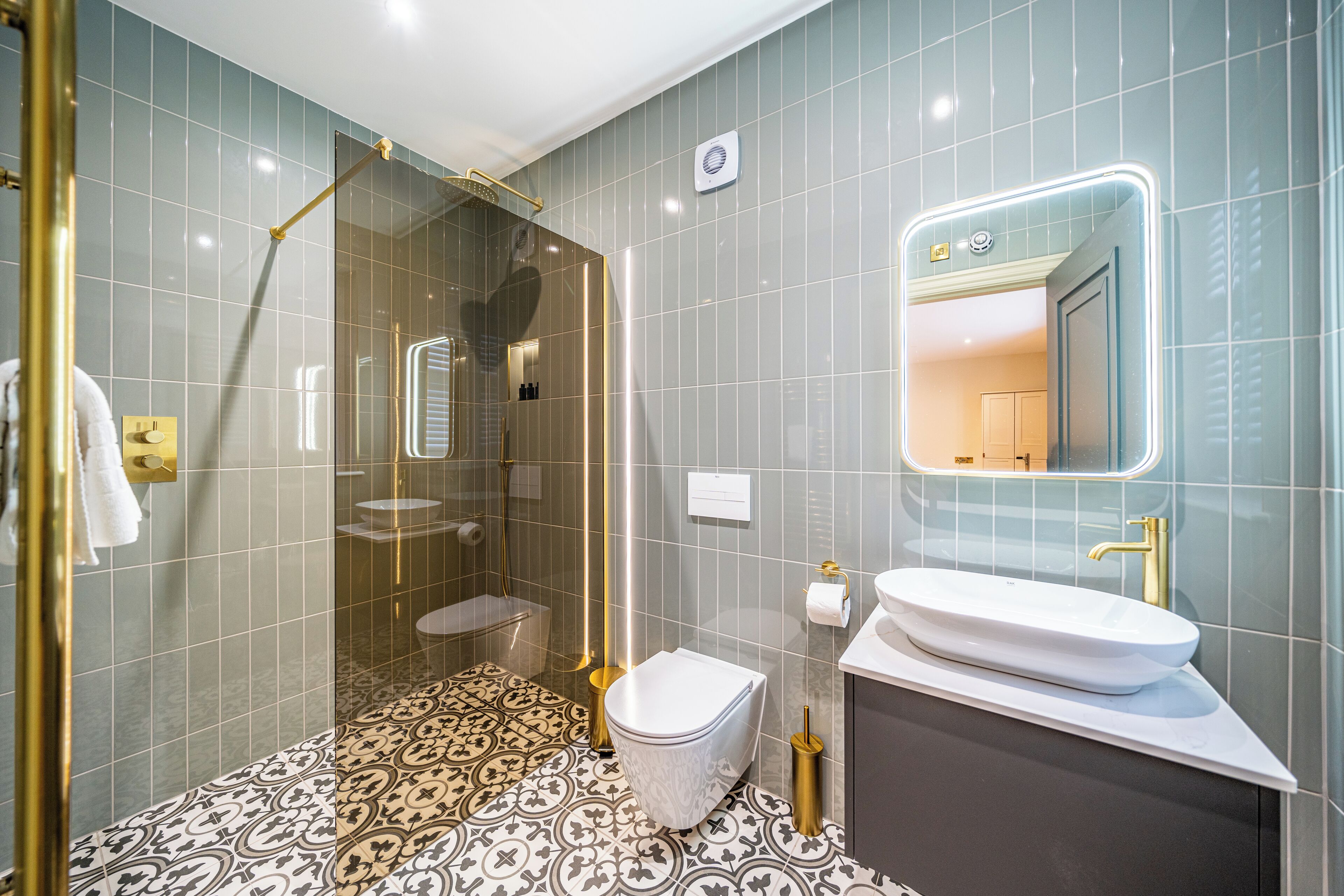 Une salle de bain moderne avec une douche à parois de verre, des toilettes blanches, un lavabo avec un robinet doré et des carreaux de sol à motifs.