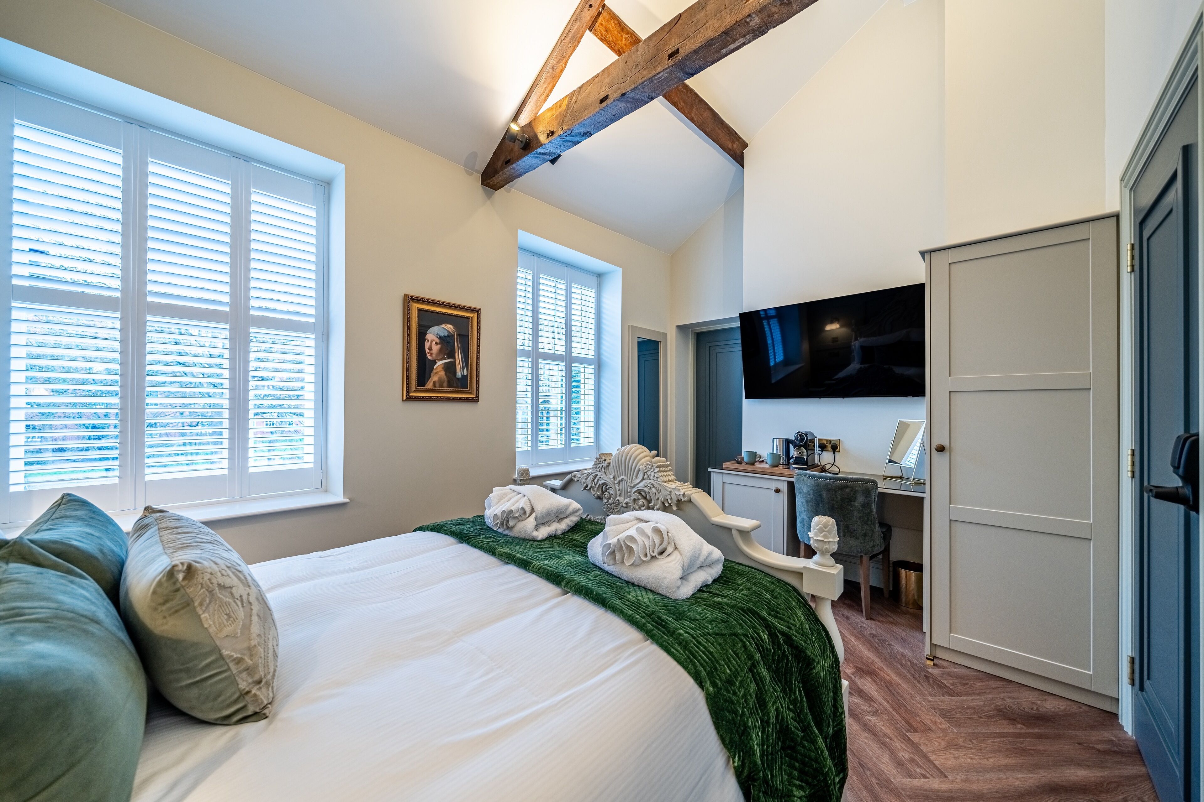 A bedroom with a large bed, two windows with blinds, a desk with a computer, and a wardrobe.