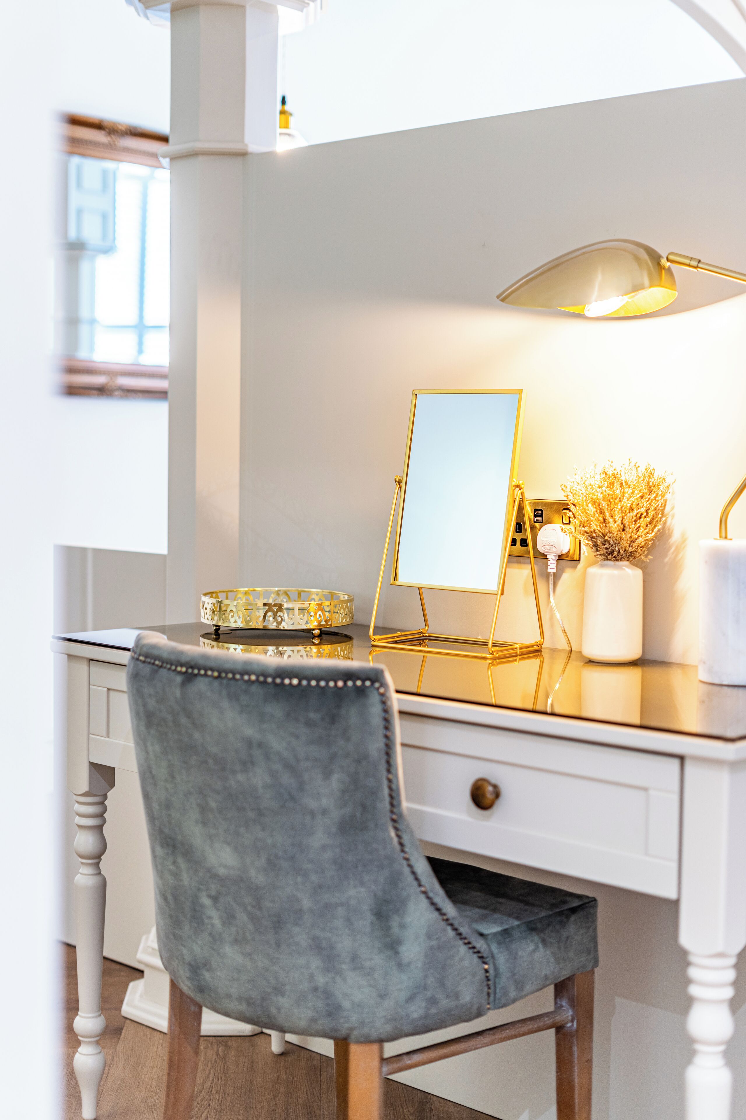 A modern desk with a gold lamp, a mirror, and a vase with dried flowers.