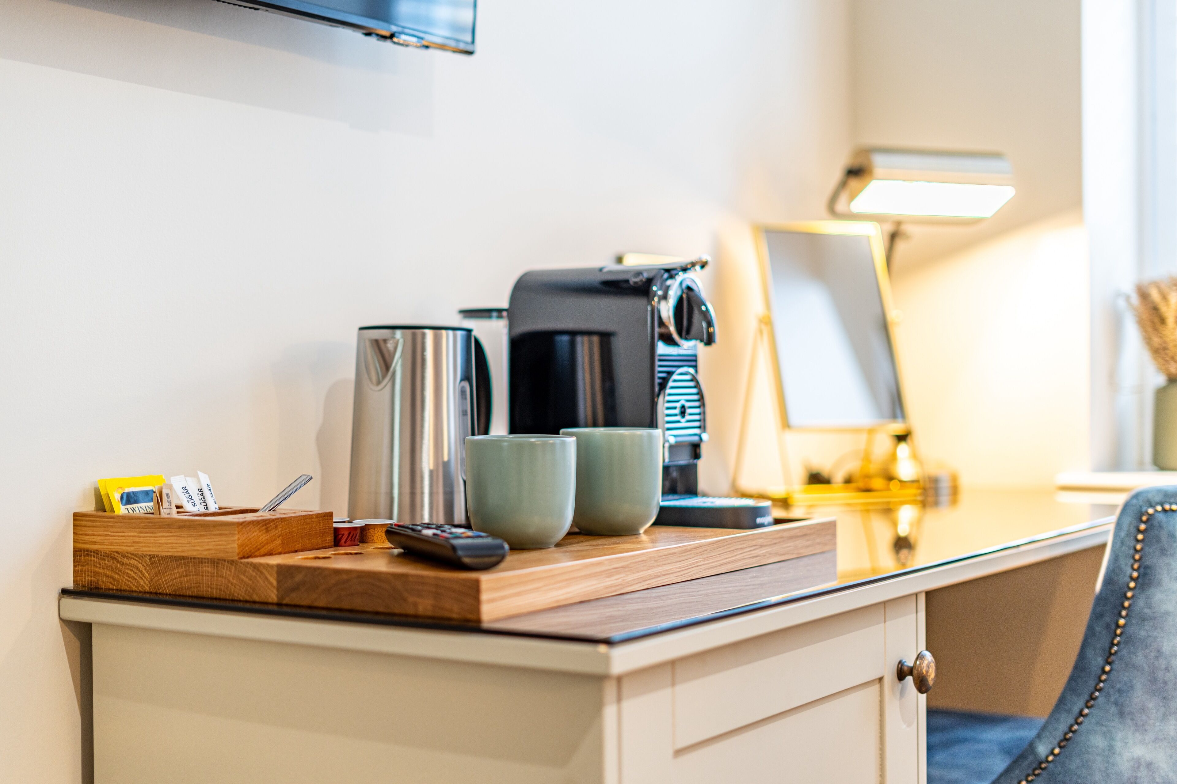 Hotelkamer met een houten bureau, een koffiezetapparaat, twee kopjes, een afstandsbediening en een lamp.