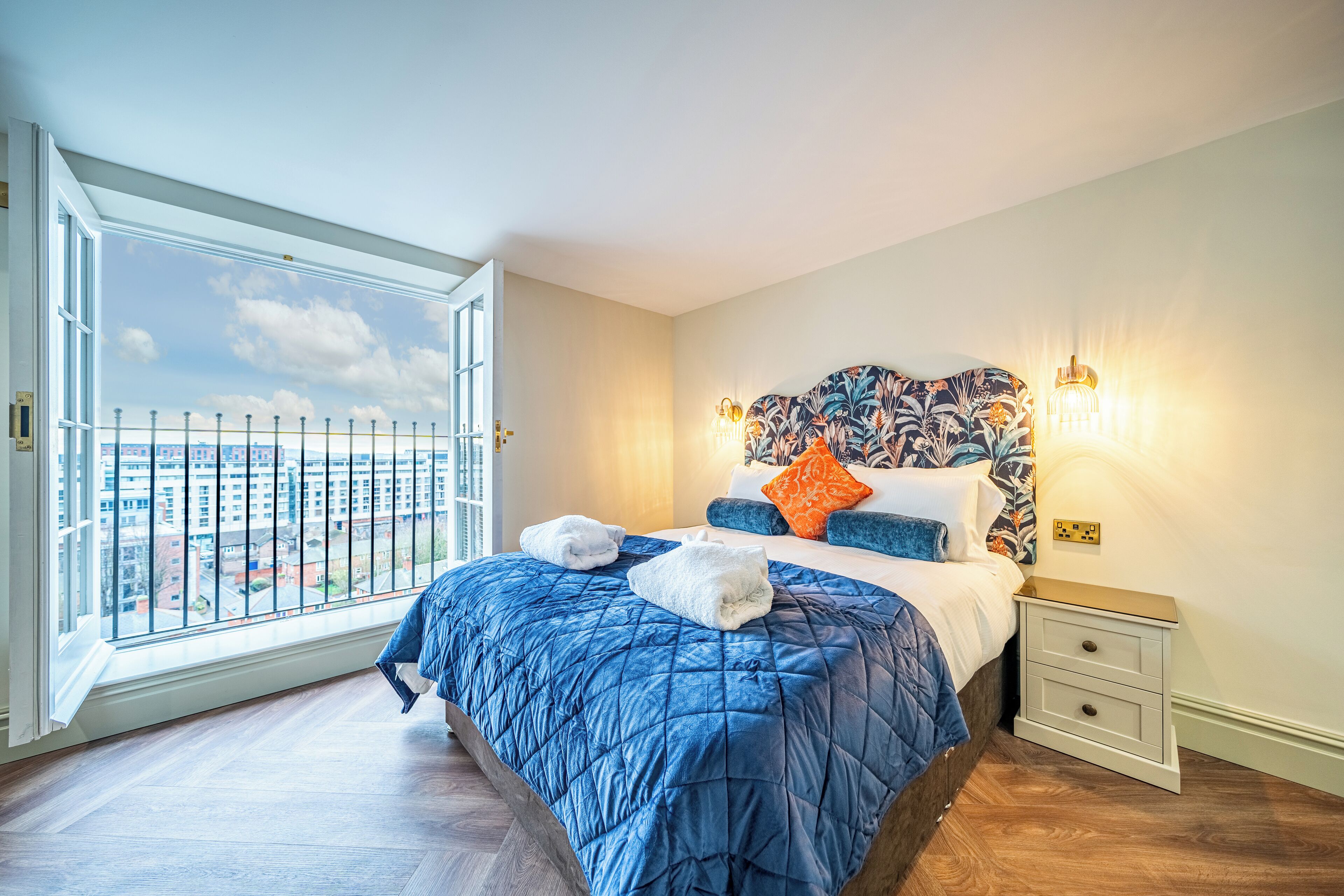 Une chambre avec une grande fenêtre, un lit recouvert d’une couette bleue, deux tables de chevet et une petite table de nuit.