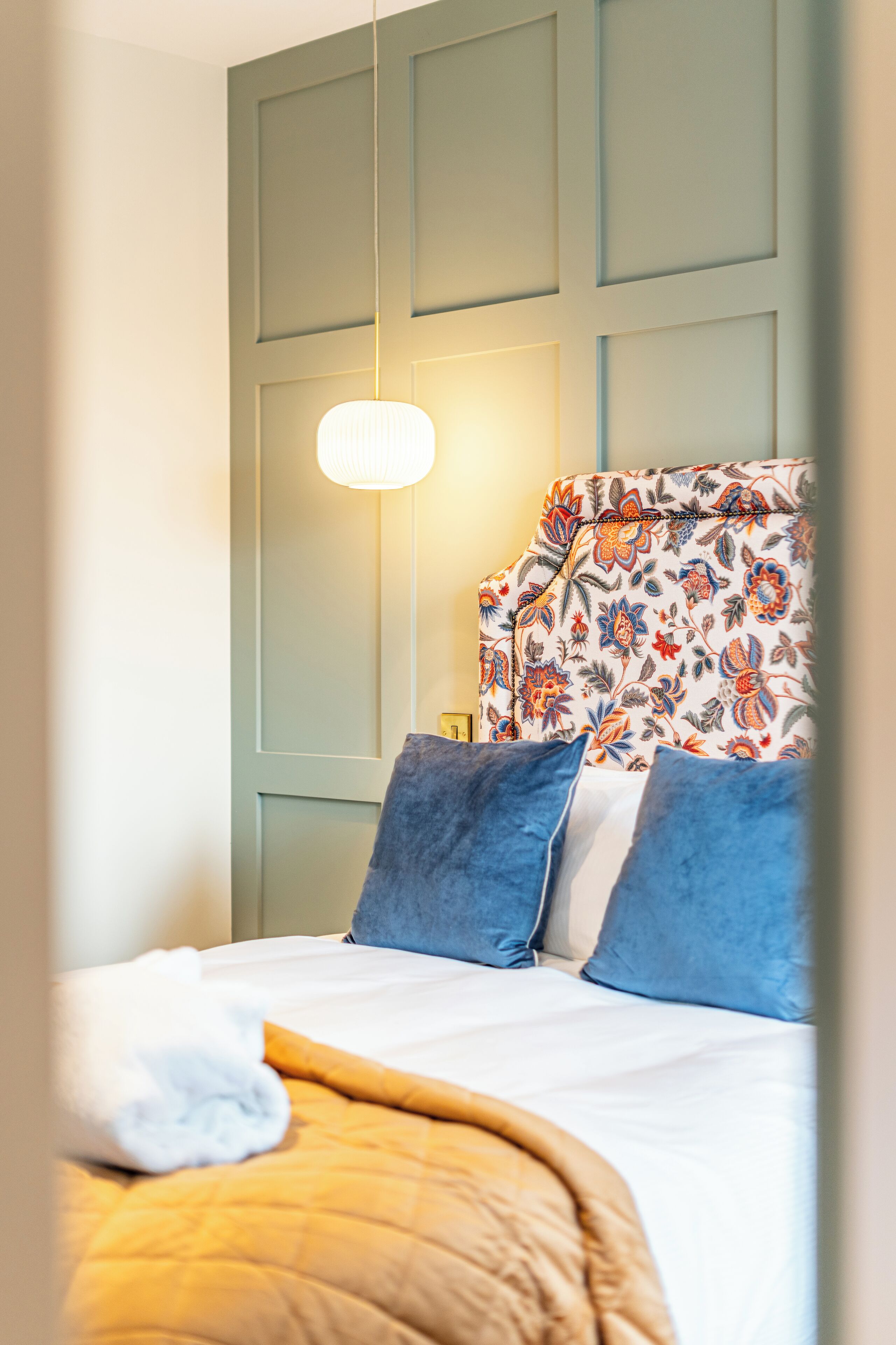 A neatly made bed with a white comforter and a brown throw blanket, a floral-patterned headboard, and two blue pillows.