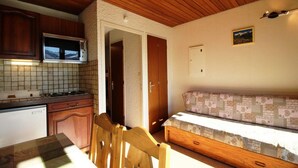 A compact kitchen with a microwave, wooden cabinets, and a tiled backsplash. There is a wooden bench with a patterned cushion and a door leading to another room.