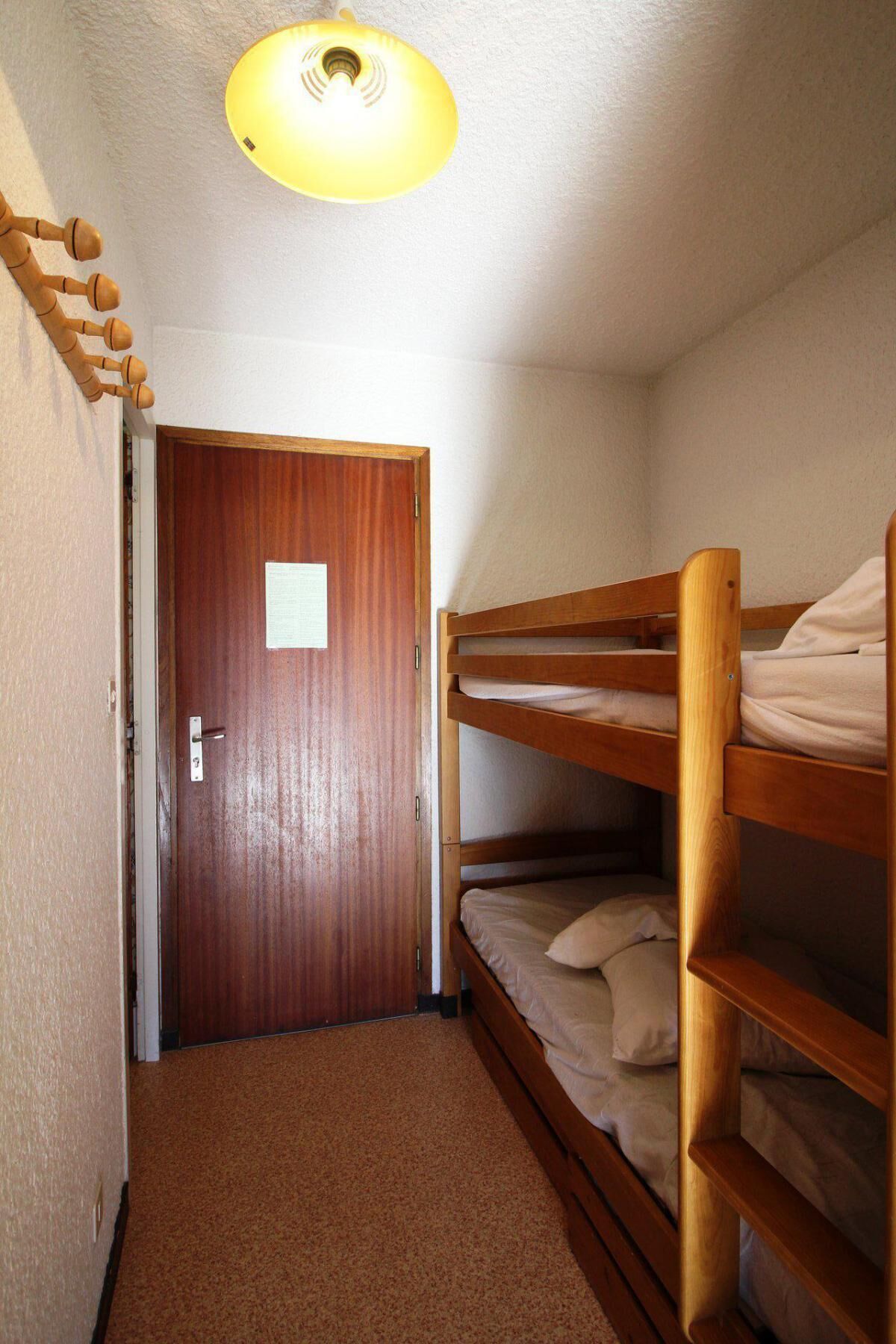 Ein kleiner, schmaler Raum mit einem hölzernen Etagenbett, einer Holztür und einer Deckenleuchte.