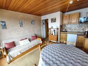 A cozy room with a bed, a sofa, a dining table, wooden cabinets, and a kitchen area with a microwave and refrigerator.