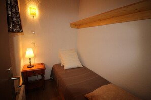A small, cozy room with a single bed, a nightstand with a lamp, and a wooden shelf above the bed. - 300m from resort center, Ground floor, view valley, balcony, tv, ski locker, 19m², Auris en Oisans (Auris)