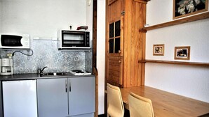 Een kleine keuken met een magnetron, fornuis en spoelbak. Er staat een houten eettafel met stoelen.