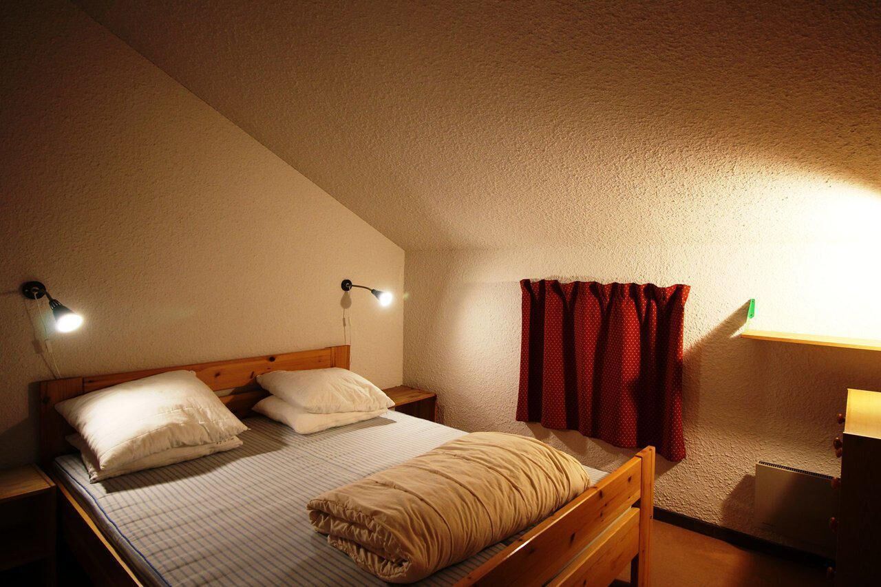 A small, cozy bedroom with a single bed, two bedside lamps, a window with red curtains, and a wooden headboard.