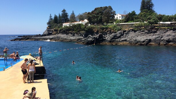 A beach with clear water, people swimming and sunbathing, and a rocky coastline with buildings in the background.