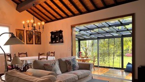 A spacious living room with a large sofa, a dining table, and a view of a lush garden through a glass door.